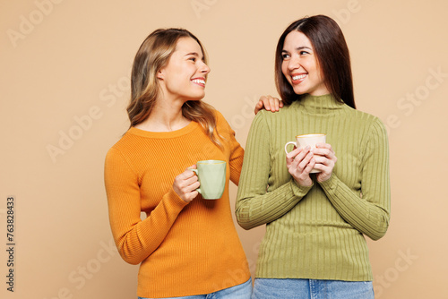 Young smiling fun friends two women they wear orange green shirt casual clothes together hold cup drink coffee look to each other isolated on plain pastel beige background studio. Lifestyle concept.