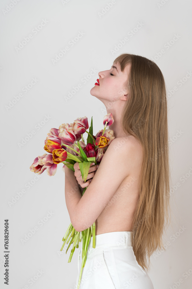 Half naked woman with her eyes closed holds bouquet of spring tulips with her hands and covers chest with flowers. Young Caucasian blonde woman with long hair. Side view