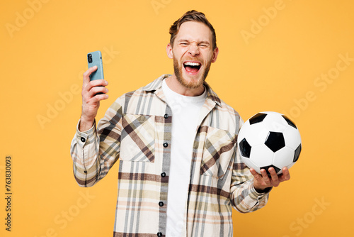 Young man fan wears brown shirt casual clothes cheer up support football sport team hold in hand soccer ball watch tv live stream use mobile cell phone scream do isolated on plain yellow background.