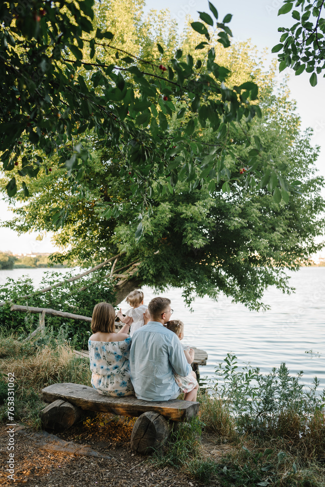 A happy family is sitting back on a bench on the shore of a lake and looking at a beautiful sunset view. Mom, dad hug children daughter near pond. Mother, father embrace kids and spend time together. A happy family is sitting back on a bench on the shore of a lake and looking at a beautiful sunset view. Mom, dad hug children daughter near pond. Mother, father embrace kids and spend time together.
