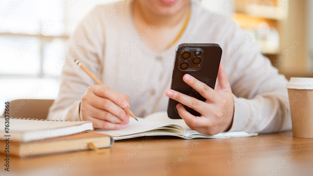 Fototapeta premium A cropped shot of a woman using her phone while working on her tasks at a table in a coffee shop.