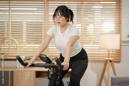 A strong Asian woman in sportswear is exercising on a fitness bike, doing indoor cardio at home. © bongkarn