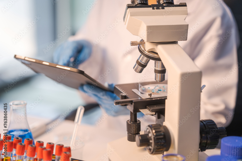Close up female scientist in white lab coat working in biotechnological laboratory, using microscope, pipette, glass flask in clinical laboratory. Close up female scientist in white lab coat working in biotechnological laboratory, using microscope, pipette, glass flask in clinical laboratory.