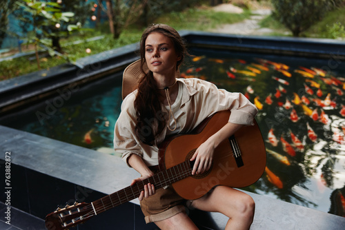 A woman playing an acoustic guitar in front of a pond with goldfish in it