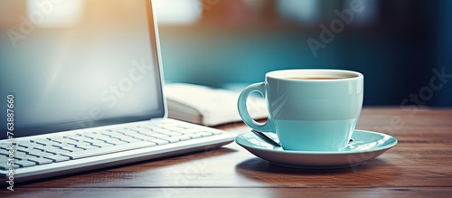 A cup of coffee sits on a saucer next to a laptop on a table