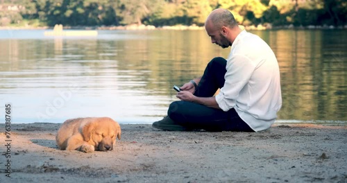 A middle-aged man sits on the beach with his faithful dog, enjoying the tranquil atmosphere of the seaside bay. Amidst the peace by the sea, a moment filled with the serenity of nature is captured