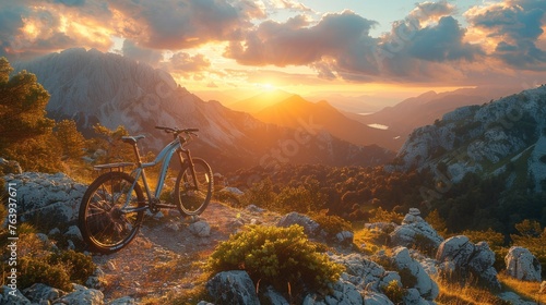 A bike sits stationary on the rocky summit of a mountain as the sun descends below the horizon, casting warm hues across the sky and landscape