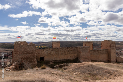 A historic stone fortress with three flags on top overlooks a vast landscape under a cloudy sky, showcasing architectural and natural beauty