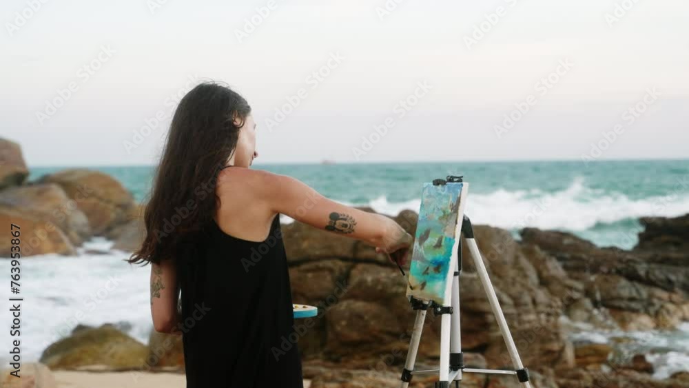 Female artist paints seascape on canvas at rocky shore. Waves crash, woman in casual dress enjoys painting hobby, captures ocean beauty. Art creation, landscape inspiration in serene nature. Slowmo Female artist paints seascape on canvas at rocky shore. Waves crash, woman in casual dress enjoys painting hobby, captures ocean beauty. Art creation, landscape inspiration in serene nature. Slowmo