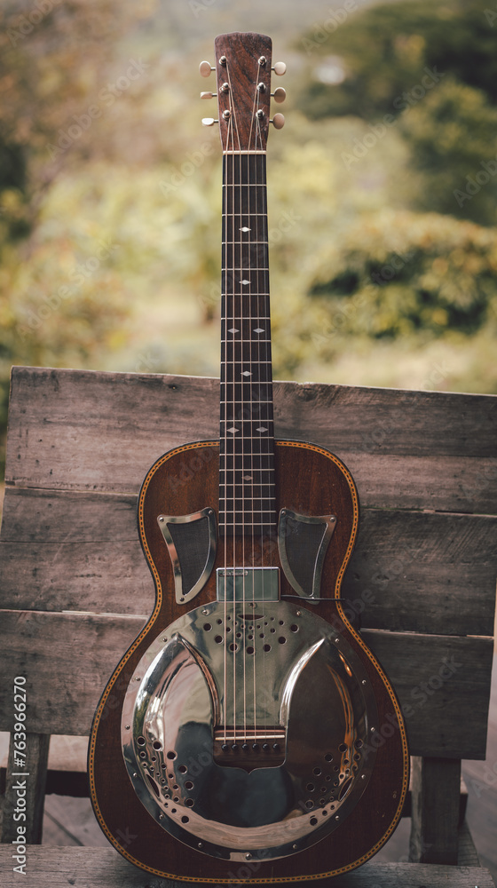 Fototapeta premium A guitar with a hat on top of it is sitting on a wooden platform. The guitar is a small, old-fashioned style, and the hat is black. The scene is set in a wooded area with mountains in the background