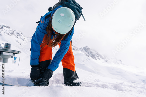 woman snowboarder fastens snowboard bindings while standing on top of  slope in ski resort. Against background of grandiosely beautiful mountain range