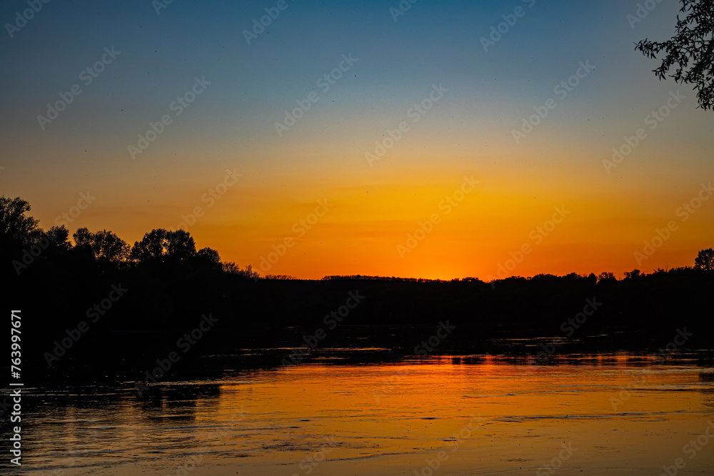 Great Don River in middle reaches. Orange sunset is reflected in water. Black silhouette of coastal forest. There are many trichopterans flying over smooth whirlpool current. Spring and high water