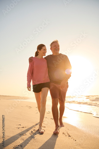 Loving Retired Senior Couple On Vacation Walking Along Beach Shoreline Hugging At Sunrise
