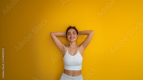 pretty young woman with hair bun, smiles pleasantly, dressed in casual outfit, keeps arms raised up behind back, isolated over yellow background copy space photo