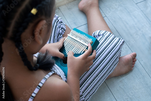 Little girl with African style braids sits and plays the kalimba thumb piano keyboard on floor, view from above photo