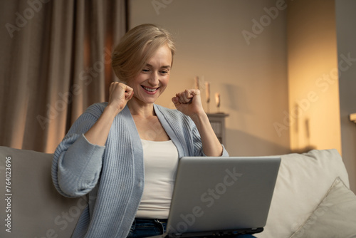 A joyful woman is seated on a sofa with a laptop on her knees, clapping her hands with an expression of elation. The warm, comfortable ambiance of the living room suggests a personal triumph