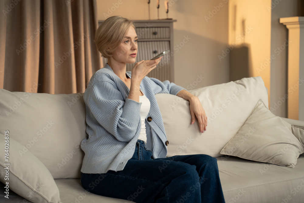 A woman is seated comfortably on a light-colored couch, holding a smartphone in front of her face as she records a voice message. She appears focused and engaged, with a serene living room setting in A woman is seated comfortably on a light-colored couch, holding a smartphone in front of her face as she records a voice message. She appears focused and engaged, with a serene living room setting in