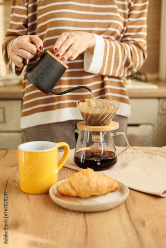 female makes coffee in morning kitchen. brews in purover. yellow cup with black coffee drip. daily routine of happy woman. Food and drinks, croissant on table. Americano cappuccino for cheerfulness