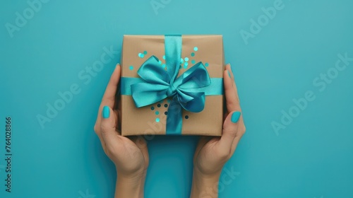 First person top view photo of hands holding craft paper giftbox with vivid blue ribbon bow over shiny sequins on isolated pastel blue background