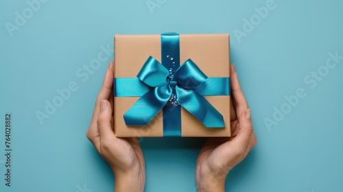 First person top view photo of hands holding craft paper giftbox with vivid blue ribbon bow over shiny sequins on isolated pastel blue background