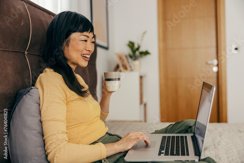 A happy japanese woman is sitting on a bed, drinking coffee and scrolling on a laptop.