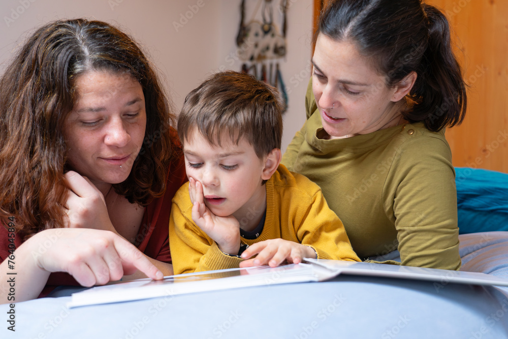 Couple of lesbian mothers watching a book with their son in the bedroom at home Couple of lesbian mothers watching a book with their son in the bedroom at home