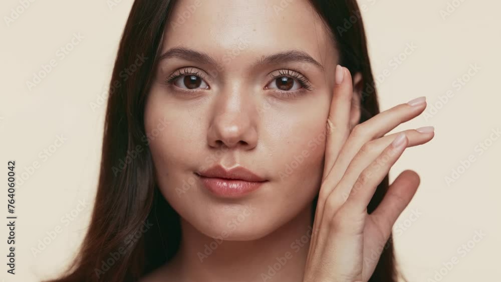 Calm young woman with a serene expression and her hand gently resting on her face against a neutral beige backdrop. Calm young woman with a serene expression and her hand gently resting on her face against a neutral beige backdrop.