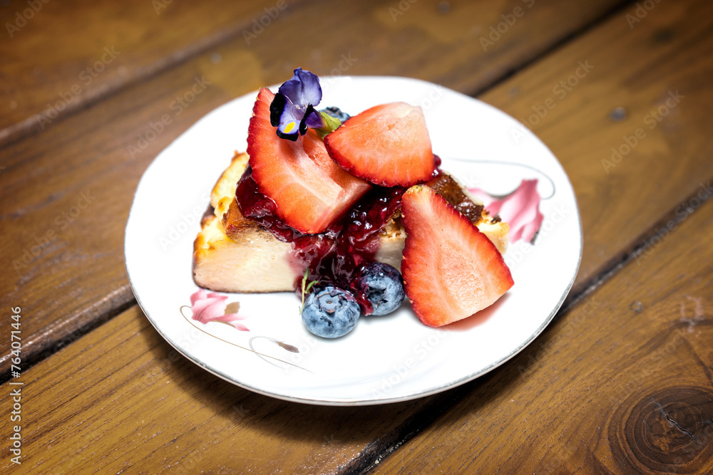 Basque burnt cheesecake topped with jam and fresh strawberries and blueberries in a plate on a vintage style wooden table in a cafe. Basque burnt cheesecake topped with jam and fresh strawberries and blueberries in a plate on a vintage style wooden table in a cafe.