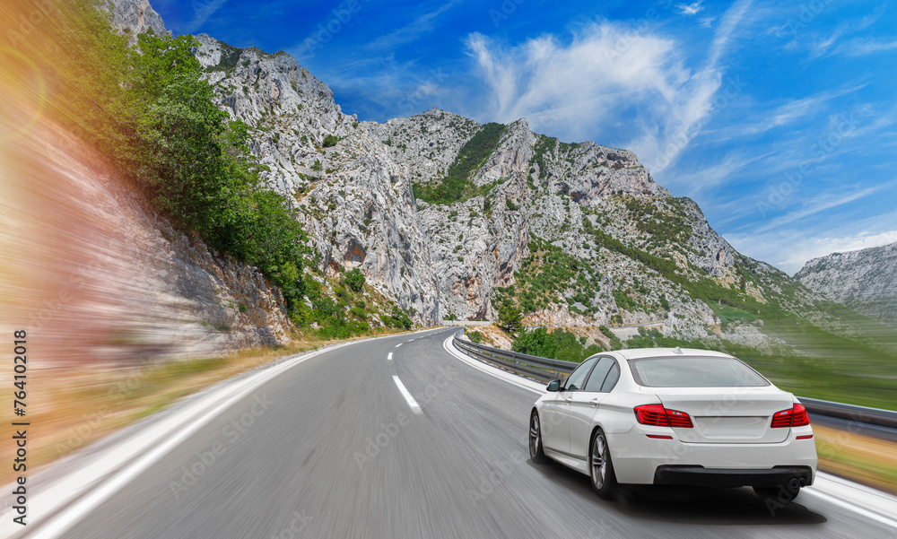 A white car drives along the highway against the backdrop of rocky mountains on a sunny day.
