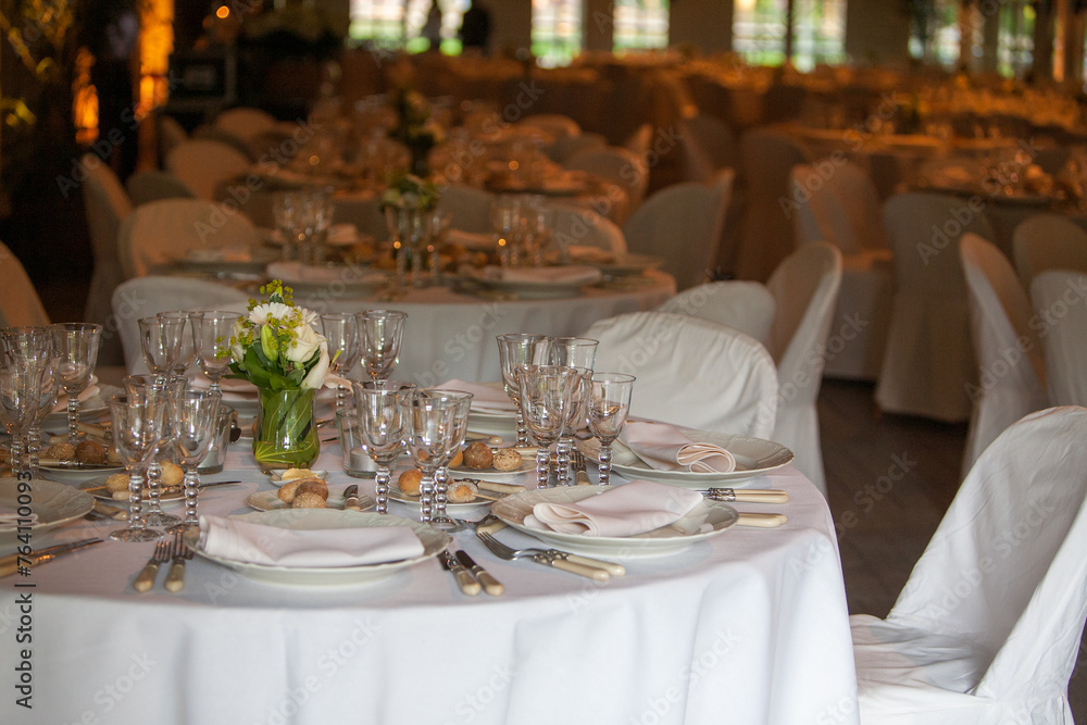 A view across a grand banquet hall adorned with multiple round tables set with white linens, sparkling glassware, and fine silverware. Each table is centered with a delicate floral arrangement