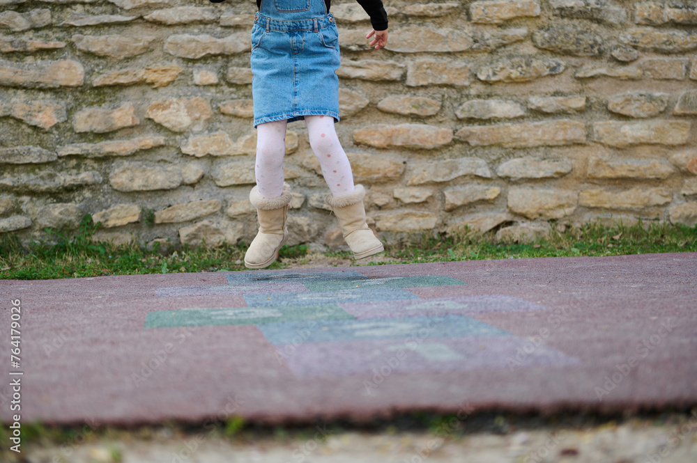 Close-up of a sporty kid girl playing hopscotch, taking turns jumping over squares marked on playground.