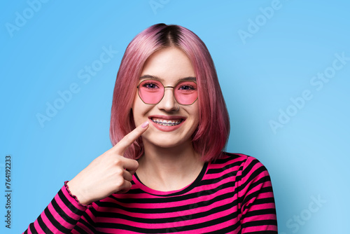 Dental dent care ad concept image - pink funny young woman girl in metal braces wear sunglasses eye glasses, stripes long sleeve shirt pointing white teeth smile. Isolated blue wall background