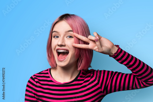 Photo image - happy pink beautiful woman in braces show v-sign victory peace hand gesture two fingers, with opened mouth, isolated blue wall background. Student girl at studio. Dental care ad concept