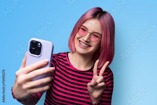 Portrait photo - pleasant pink woman in braces wear sunglasses eye glasses making taking selfie or show her viewer positive result with cell phone smartphone, v-sign gesture isolated blue background