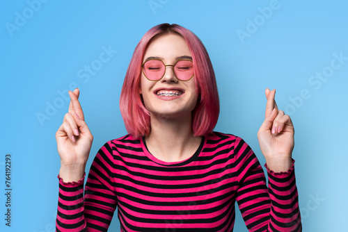 Wishful positive hopeful smiling woman student schoolgirl wear sunglasses glasses spectacles braces, crossing fingers, close eyes praying and hoping for good luck, isolated blue wall background