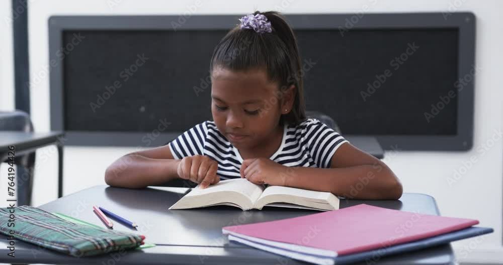 In school, in a classroom, a young African American girl is focused on reading a book In school, in a classroom, a young African American girl is focused on reading a book