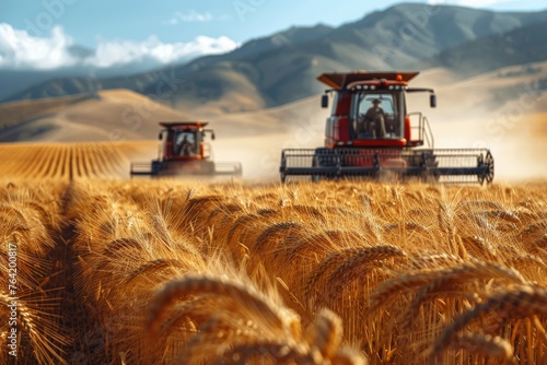 Two red harvesters kicking up dust while reaping wheat in a picturesque golden field against a backdrop of hills photo