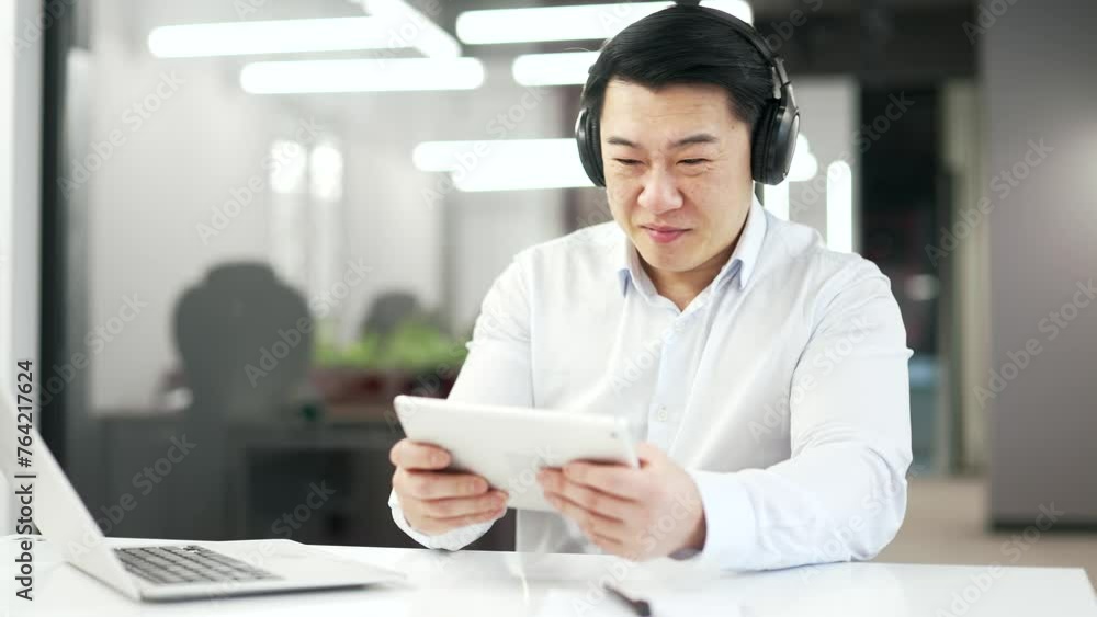 Happy asian man in wireless headphones plays a computer game on a digital tablet while sitting at desk at workplace in business office. Excited smiling mature worker engrossed in interesting game Happy asian man in wireless headphones plays a computer game on a digital tablet while sitting at desk at workplace in business office. Excited smiling mature worker engrossed in interesting game