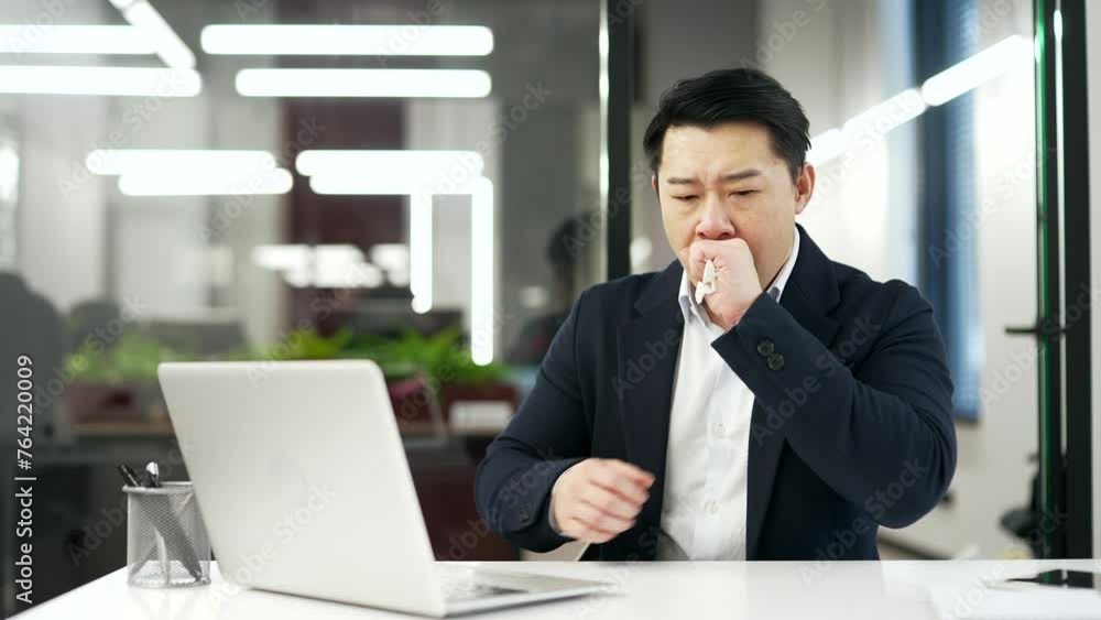 Asian businessman coughing while working on laptop sitting at workplace in business office. Sick with a cold or flu mature man feels bad, holds his hand to his chest, blows his nose with handkerchief Asian businessman coughing while working on laptop sitting at workplace in business office. Sick with a cold or flu mature man feels bad, holds his hand to his chest, blows his nose with handkerchief
