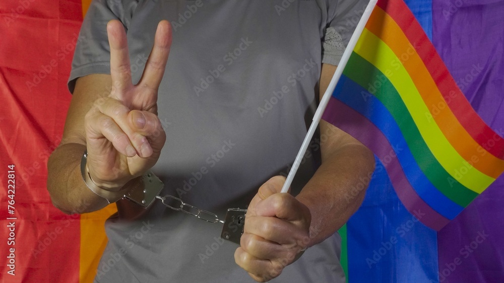 Obraz premium The man holds the rainbow flag and with the other hand makes the victory sign at the camera despite having handcuffs on his wrists