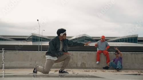 Handsome break dancer enjoy perform freestyle movement with asian friend cheering behind. Hipster listen to music from headphone and looking at camera. Street dance. Outdoor sport 2024. Sprightly.