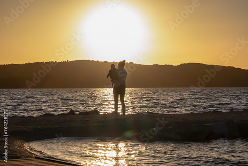 Silhouette of loving mother holding small toddler at romantic sunset in Okrug Gornij, Split-Dalmatia, Croatia. Coastline of island Otok Ciovo, Adriatic Sea in summer. Family travel vacation concept photo