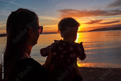 Mother with small child enjoying romantic sunset at beach in town Makarska, Split-Dalmatia, Croatia, Europe. Coastline of Makarska Riviera, Adriatic Sea. Dreamlike atmosphere. Family vacation concept photo