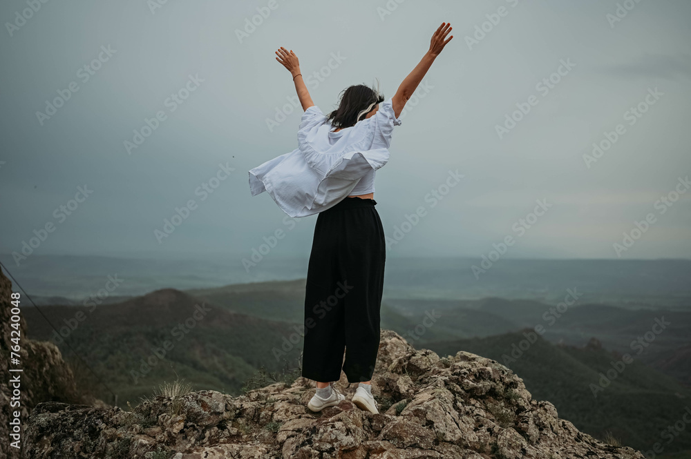 Happy woman tourist hiking up mountain enjoying nature. Landscape in cloudy gloomy windy weather. Female in white shirt with open arms outstretched in joy, enjoying travel. Freedom concept Happy woman tourist hiking up mountain enjoying nature. Landscape in cloudy gloomy windy weather. Female in white shirt with open arms outstretched in joy, enjoying travel. Freedom concept