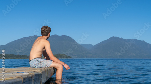 Teenage boy in swimwear sitting on the lake dock  gazing at the horizon with mountains in the background. Perfect summer snapshot.