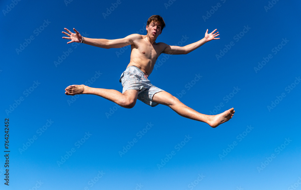 A joyful young man leaps in the air with arms wide open and a surprised expression, against a backdrop of beautiful blue sky on a sunny summer day.