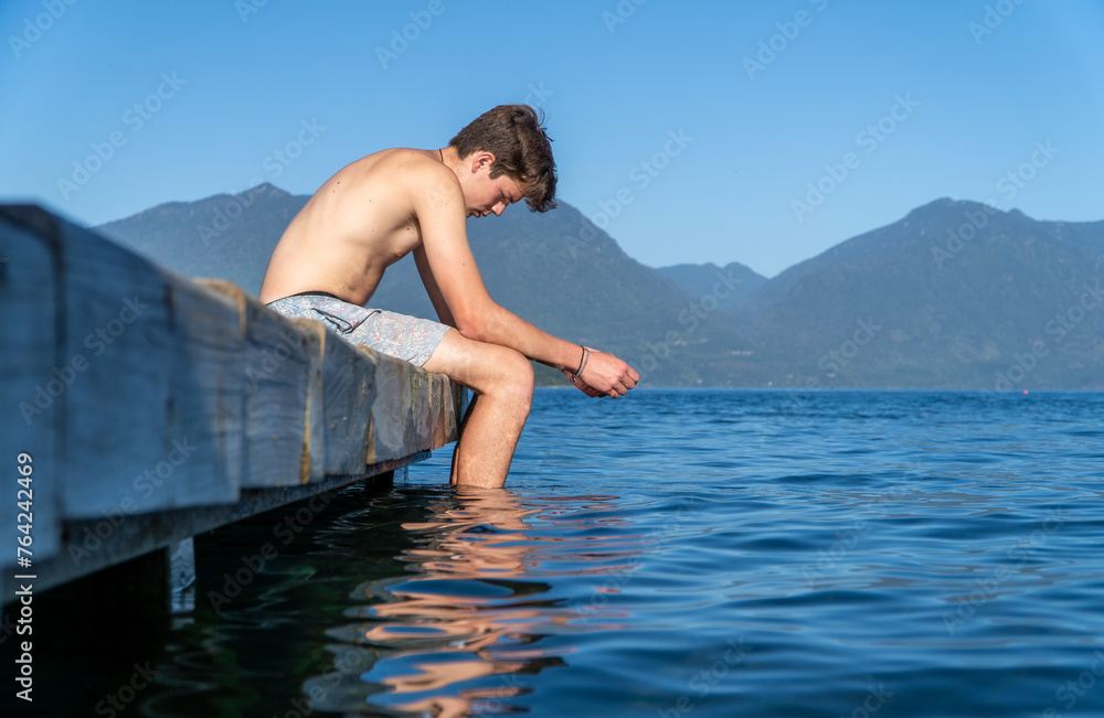 Young man sitting at the edge of the dock, looking down with a sad and worried expression, feet dangling in the water on a beautiful summer day, depicting teenage depression and solitude.