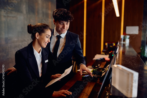 Happy receptionists cooperating while working on computer at hotel front desk.