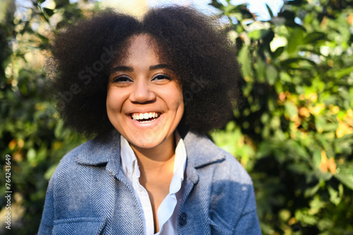 Fashion close up stylish portrait of attractive young natural beauty African American woman with afro hair in blue coat and white shirt posing outdoors. Happy lady laugh with perfect smile and teeth