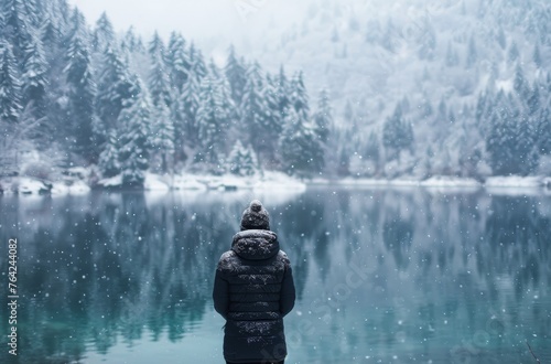 A person is standing in the snow near a lake. The person is wearing a black coat and he is looking out over the water. The scene is peaceful and serene